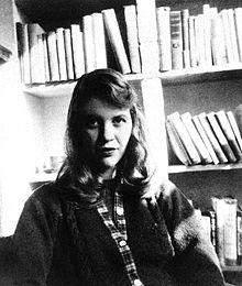 A black-and-white photo of a woman with shoulder-length hair. She is seated facing the camera, wearing a sweater, with bookshelves behind her.
